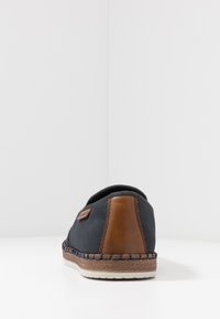 Chaussure en toile sombre avec un accent en cuir marron au talon, des détails cousus et une semelle tissée ressemblant à de la paille. Design simple à enfiler sans lacets visibles.