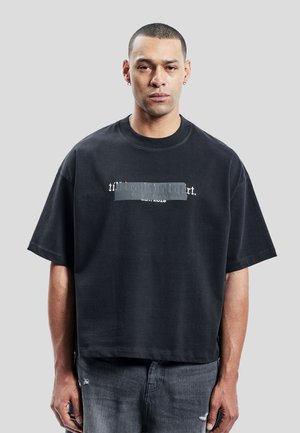 Mand med kort hår iført en sort oversized t-shirt med delvist skjult hvid tekst og falmede sorte jeans mod en grå baggrund.