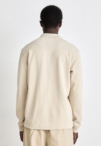 Camisa de manga longa bege com colarinho, feita de tecido texturizado, apresentando um design limpo e minimalista com um ajuste descontraído.