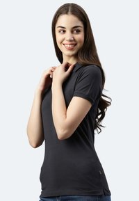 Schwarzes Polo-Shirt mit kurzen Ärmeln, glatter Textur, taillierter Passform und einem kleinen Etikett am Saum. Getragen mit blauen Jeans.