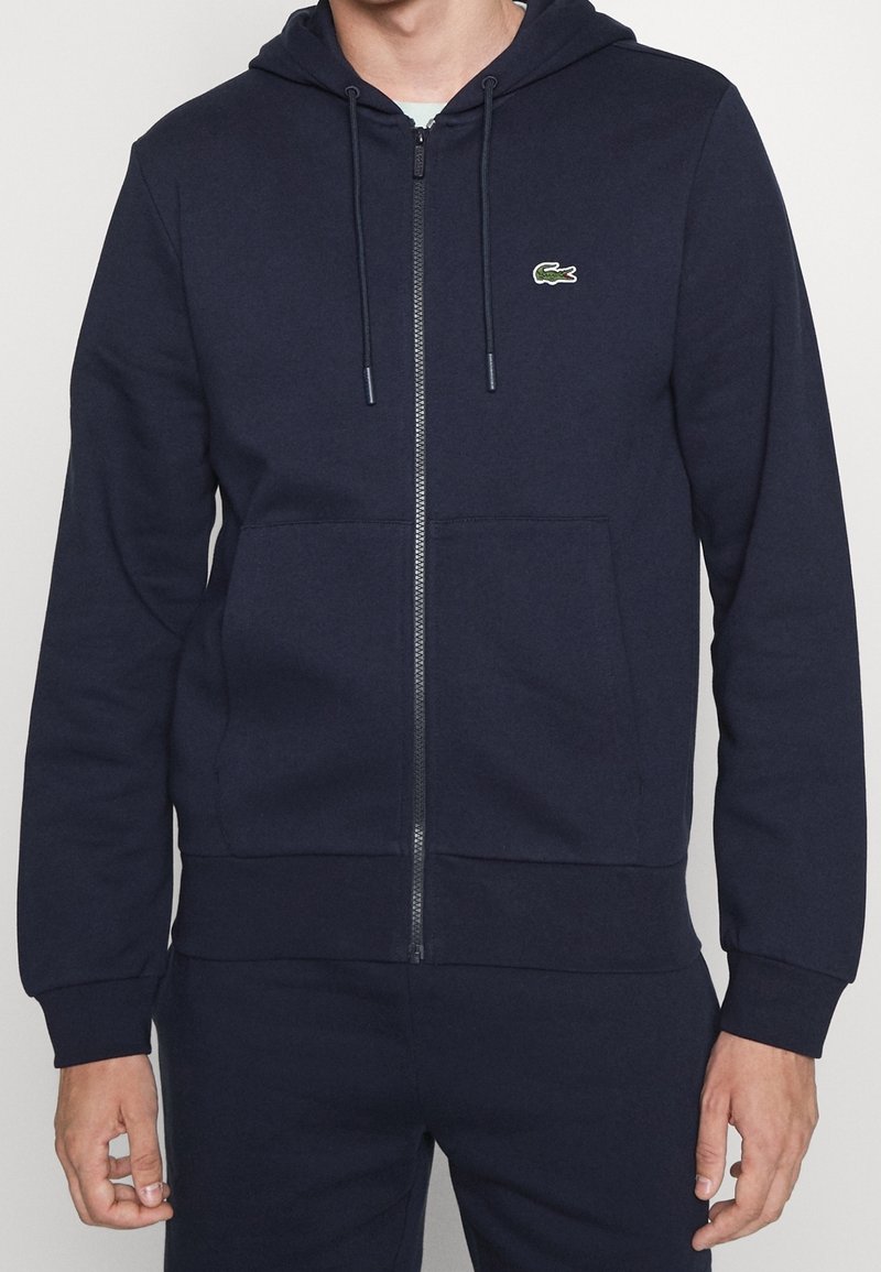 Homme portant un sweat à capuche zippé bleu marine avec des cordons et des poches avant, avec un petit logo crocodile vert sur la poitrine gauche.