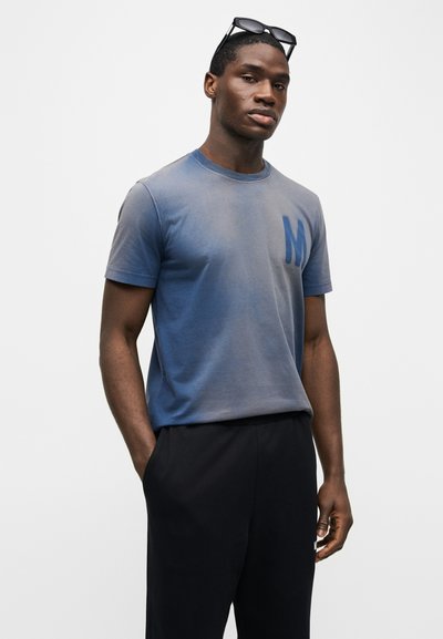 Άνδρας που φοράει ξεβαμμένο μπλε μπλουζάκι με λογότυπο "M", μαύρο παντελόνι και γυαλιά ηλίου που ακουμπούν στο κεφάλι του, στέκεται μπροστά σε λευκό φόντο.