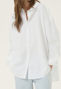 Woman wearing oversized white button-up shirt with long sleeves and light blue jeans, hands in pockets, against plain background.