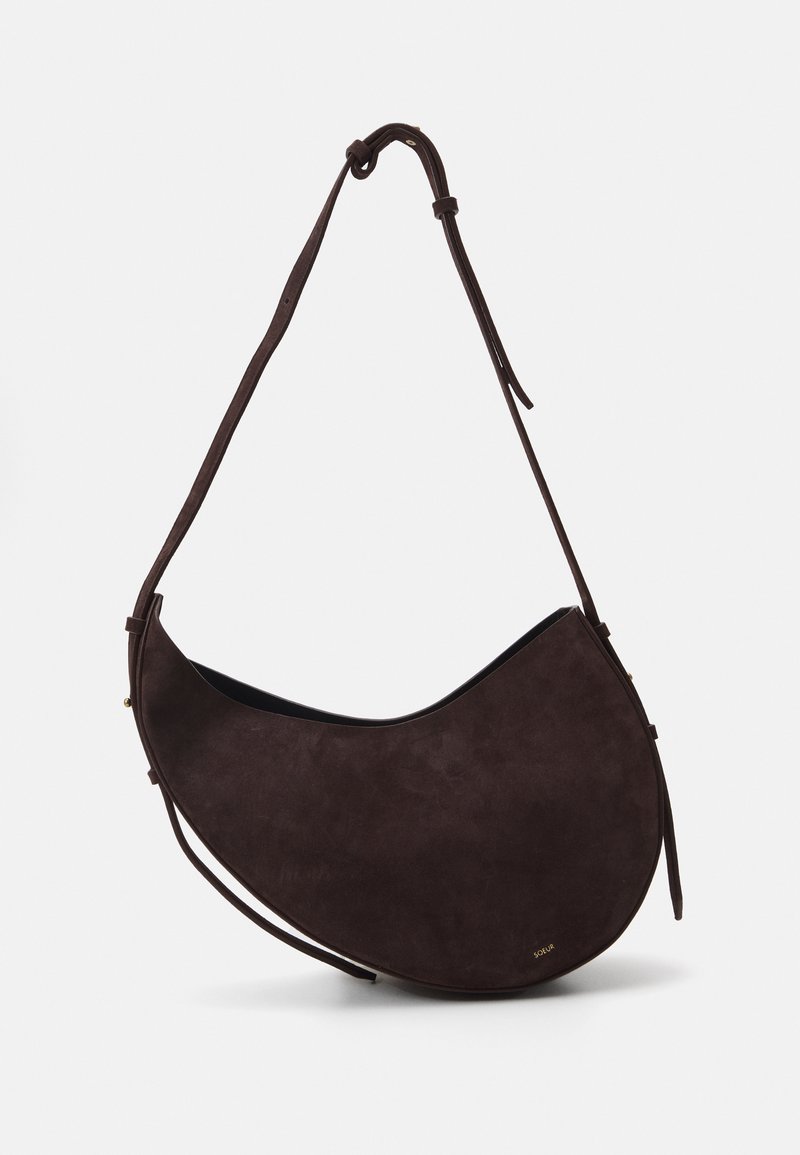 Bolso de hombro de ante marrón con una forma curvada, correa delgada ajustable y detalles de hardware mínimos. Textura suave con una apariencia delicada.