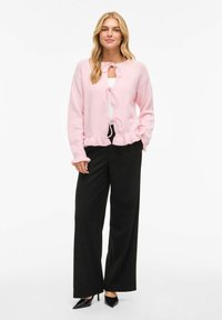 Rosa strikkejakke med volangkant, tre knytelukkinger og lange ermer, kombinert med svarte vide bukser og svarte høye hæler.