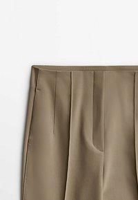 Gros plan sur un pantalon beige taille haute avec des coutures verticales et des poches latérales, présenté à plat sur un fond blanc.