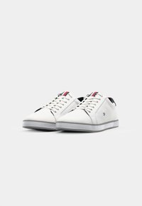 Witte canvas sneakers met een zwart en rood accent op de tong. Heeft een low-top ontwerp, platte rubberen zool en metalen oogjes voor de veters.