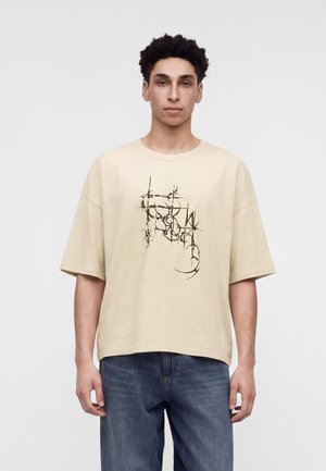 Ung mand iført oversized beige T-shirt med abstrakt sort grafik og blå jeans, stående mod en ensfarvet grå baggrund.