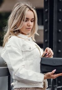 Černá kožená peněženka s klopou a zipem, držena v ruce. Osoba má na sobě bílou denimovou bundu s knoflíky a bočními kapsami.