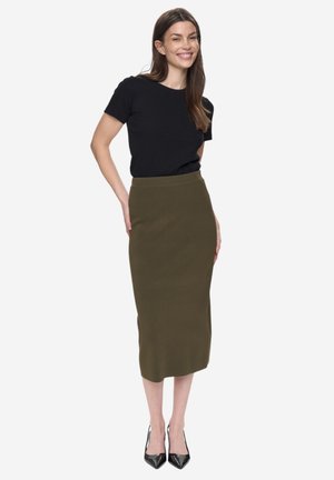 Schwarzes kurzärmeliges Oberteil kombiniert mit einem figurbetonten olivgrünen Midirock. Modell trägt schwarze spitz zulaufende High Heels. Glatter Stoff, minimalistisches Design.