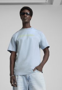 Lyseblå, kortærmet t-shirt med ribbet rund hals og grafisk tekst i neongrøn. Båret med slidte denimshorts og solbriller.