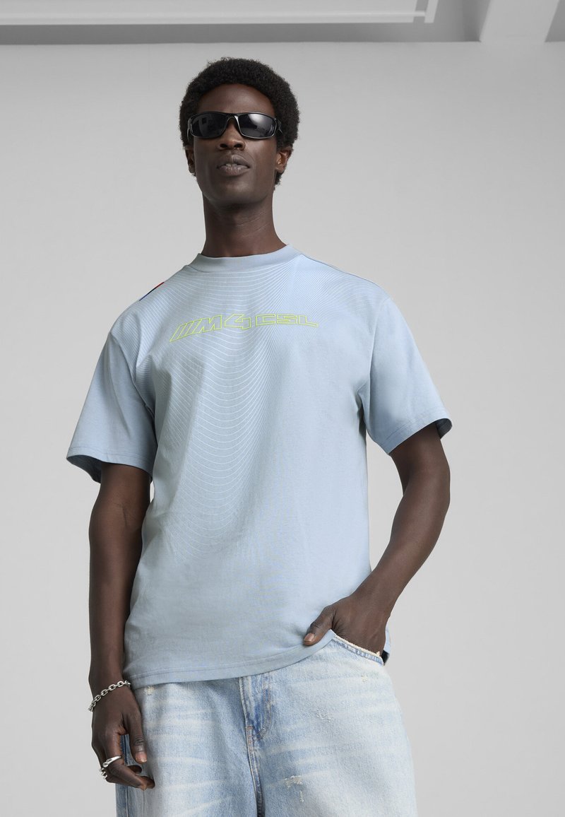 Lyseblå, kortærmet t-shirt med ribbet rund hals og grafisk tekst i neongrøn. Båret med slidte denimshorts og solbriller.
