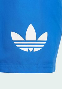 Μπλε βαμβακερές βερμούδες με ένα μεγάλο λευκό λογότυπο τριφύλλι και τρεις οριζόντιες ρίγες από κάτω. Λείο υφασμάτινο υλικό και χαλαρή σχεδίαση.