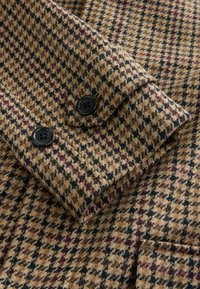 Marmurowy płaszcz wełniany w brązowo-czarny wzór houndstooth, z rękawem zakończonym dwoma czarnymi guzikami na mankiecie i widocznym stębnowaniem kieszeni z przodu.