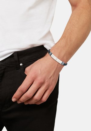 Bracciale intrecciato blu con accento argento, dotato di una piastra metallica piatta incisa con la parola "BRACCIALE", indossato su un polso accanto a jeans neri.