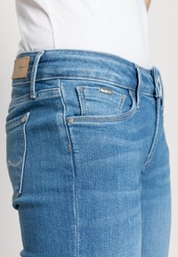Jeans de ganga azul slim-fit com lavagem clara, cinco bolsos, rebites de metal e um patch de marca em couro na cintura.