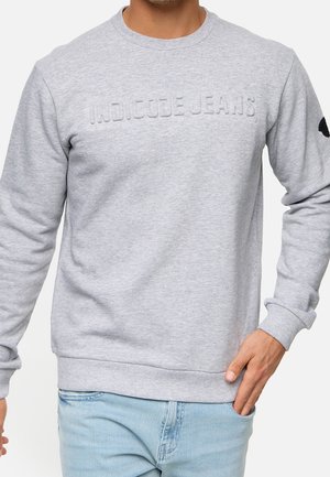 Mężczyzna w jasnoszarej bluzie z wytłoczonym napisem "INDICODE JEANS" i niebieskich jeansach, prawa ręka dotyka kieszeni.