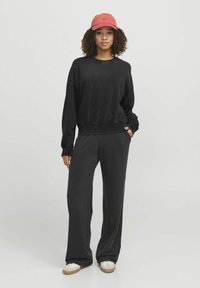 Sudadera negra de gran tamaño con puños acanalados, combinada con pantalones anchos negros. Una gorra roja se encuentra sobre el cabello, con zapatillas beige y blancas.