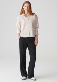 Helles, strukturiertes Cremehemd mit Knopfverschluss und hochgerollten Ärmeln, kombiniert mit maßgeschneiderten schwarzen Hosen und weißen Schuhen. Schlichte, moderne Kleidung.