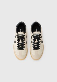 Beige sneakers with black accents and laces. Features a textured upper, rounded toe, and a gum sole. Branding visible on the tongue.