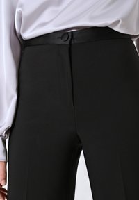 Schwarze Hose aus glattem Stoff, mit einem satinartigen Bund und einer eleganten, maßgeschneiderten Passform. Zusätzliche Details umfassen Seitentaschen.