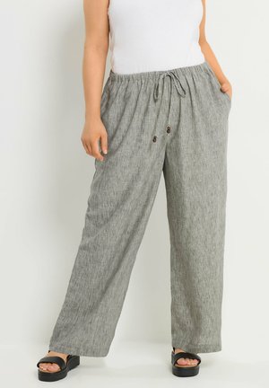 Kobieta w luźnych szarych lnianych spodniach z sznurkiem w talii i czarnych sandałach na platformie, stoi z jedną ręką w kieszeni.