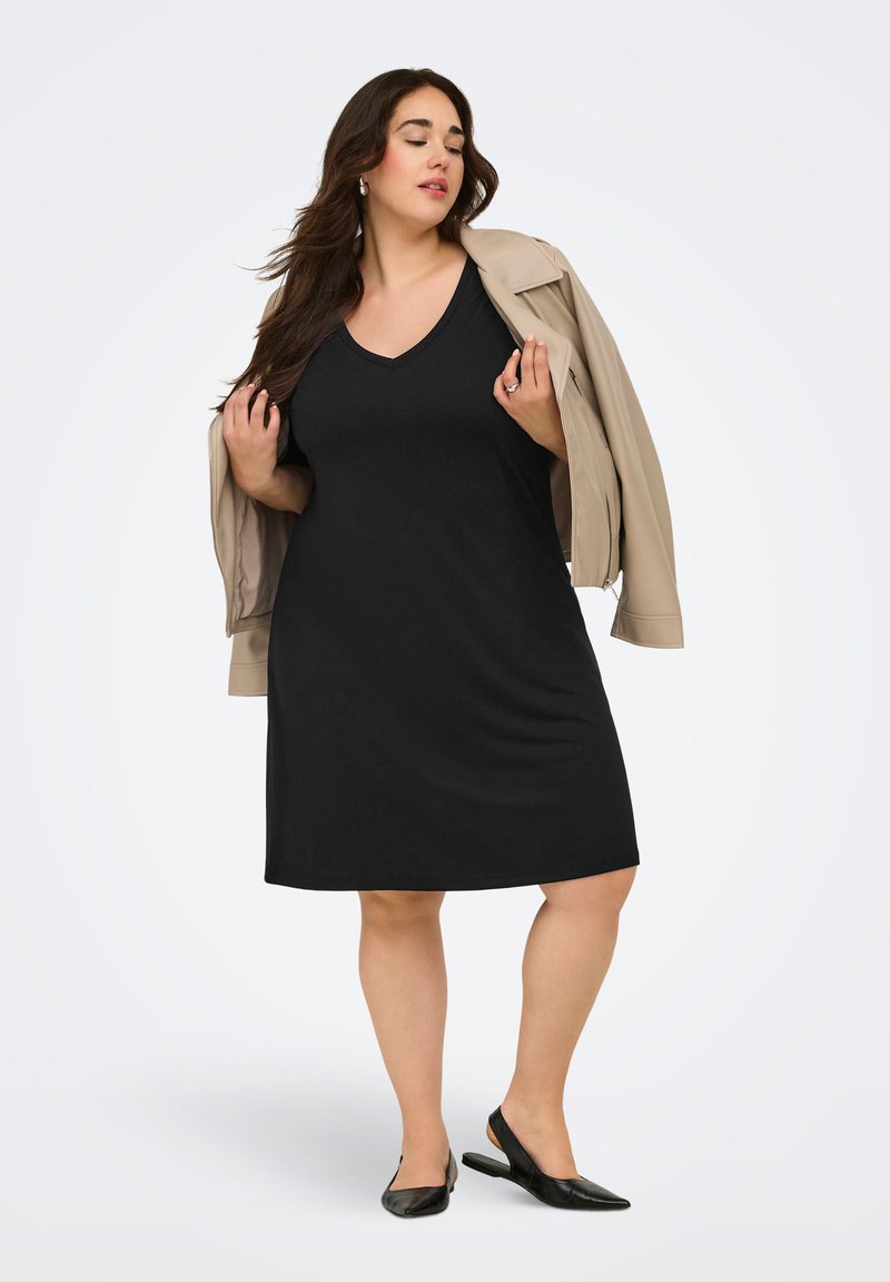 Femme portant une robe noire sans manches et une veste beige drapée sur les épaules, debout les yeux fermés devant un fond uni.