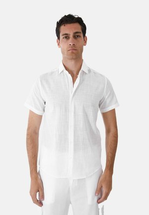 Uomo in piedi di fronte, che indossa una camicia bianca a maniche corte con bottoni e pantaloni bianchi su uno sfondo semplice.