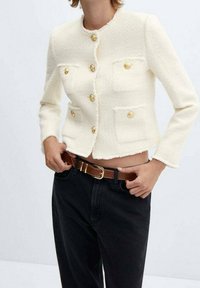 Veste courte blanche en tissu texturé, avec quatre poches avant et des boutons dorés. Associée à un jean noir et une ceinture marron.