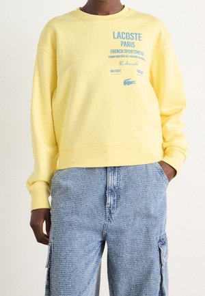 Henkilö pukeutunut keltaiseen Lacoste Paris -collegepaitaan, jossa on sininen teksti ja logo, yhdistetty vaaleansinisiin teksturoituihin cargo-housuihin, kädet taskuissa.