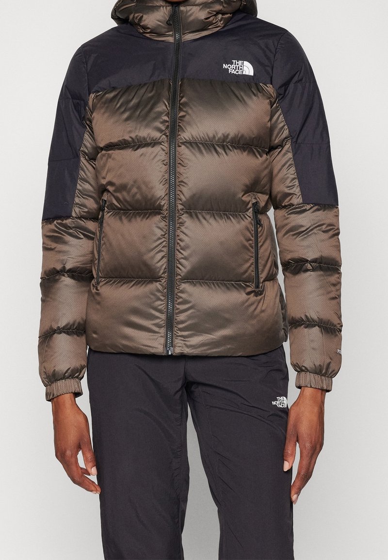 Personne portant une veste matelassée North Face bicolore marron et noire avec des poches zippées et un pantalon noir avec un logo sur la cuisse.