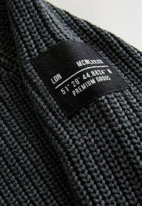 Tessuto lavorato a maglia di colore grigio scuro con motivo testurizzato. Etichetta nera attaccata con testo: "LDN MCMXLI 51° 28' 44.8824'' N PREMIUM GOODS."