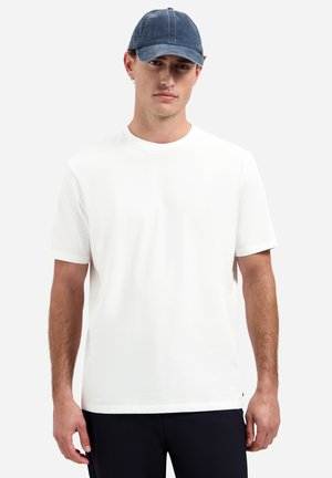 Jonge man die een eenvoudig wit T-shirt met korte mouwen, marineblauwe broek en een blauwe baseballpet draagt, staand tegen een witte achtergrond.