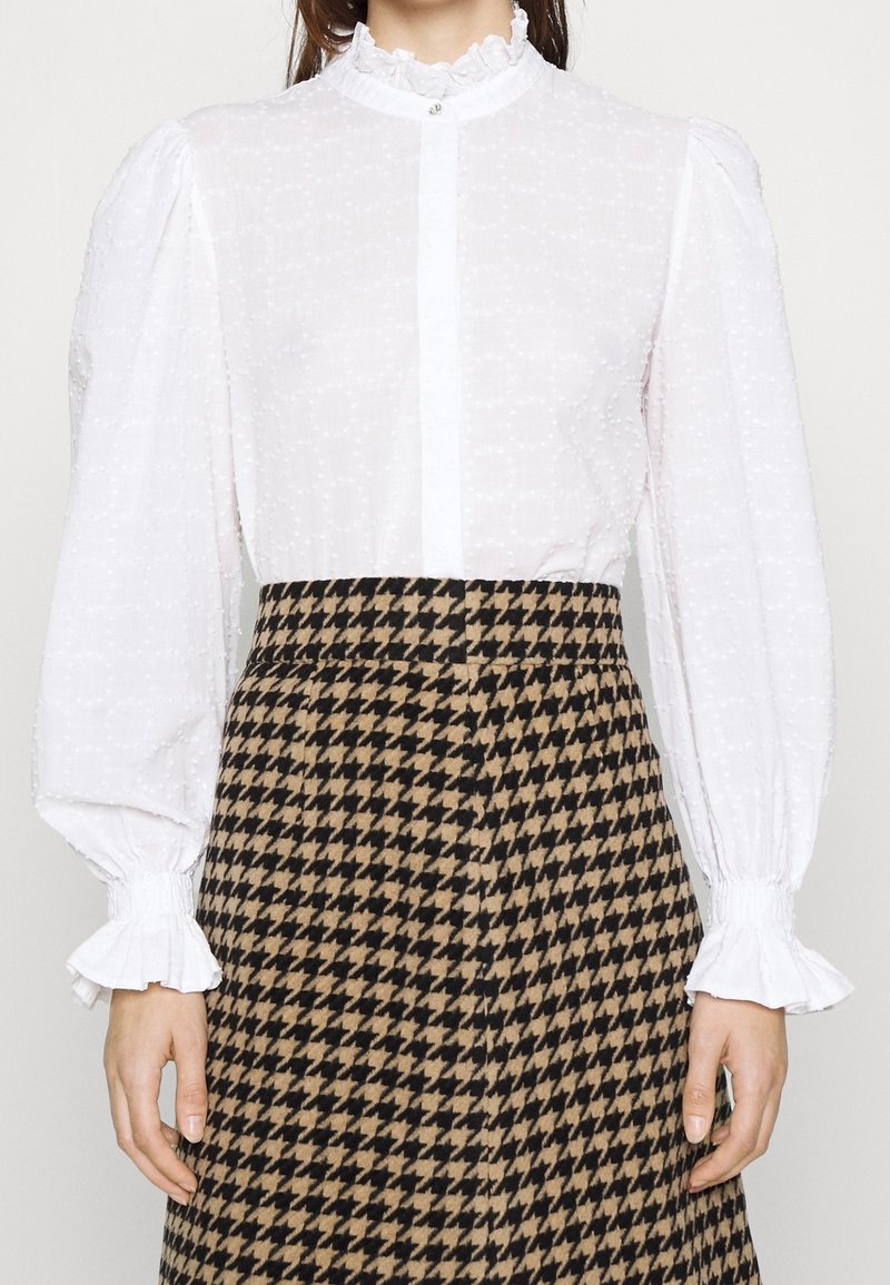 Femme portant un chemisier blanc texturé avec un col et des poignets à volants, associé à une jupe taille haute marron et noire à motif pied-de-poule.