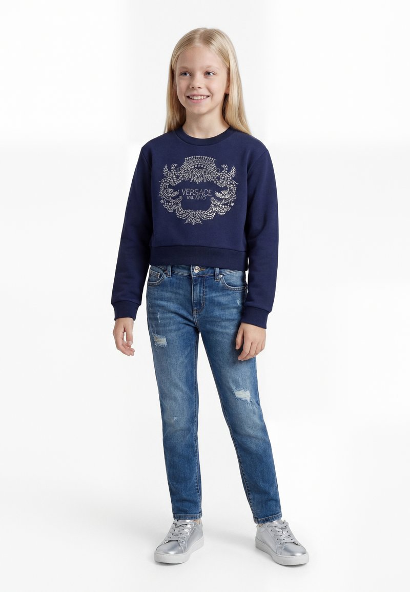 Fiatal lány mosolyog, sötétkék Versace pulóvert, kopott kék farmert és ezüst tornacipőt visel, fehér háttér előtt áll.