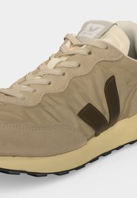 Baskets Veja beige et marron clair avec tige en daim et tissu, lacets beiges, col rembourré et semelle en caoutchouc noir, inclinées pour montrer le logo sur le côté.