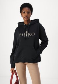 Zwarte hoodie met een voorzak en het "PINKO AELR" logo in verouderde metallic print. Er worden beige aansluitende broek gedragen.