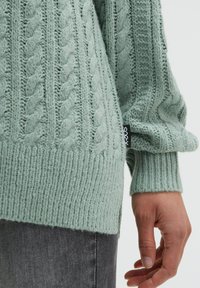 Hellgrüner Strickpullover mit Zopfmuster, gerippten Bündchen und Saum, ergänzt durch ein schwarzes Markenschild an der Seite.