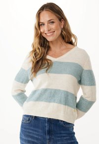 Lächelnde Frau mit langen, welligen Haaren, die einen cremefarbenen und hellblauen gestreiften Pullover mit subtilen Glitzerakzenten und blaue Jeans trägt, Hände hinter dem Rücken.