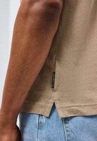 Κοντινή λήψη του μπράτσου ενός ατόμου που φοράει μπεζ πουκάμισο με μικρή μαύρη ετικέτα και ανοιχτόχρωμο μπλε τζιν, που αναδεικνύει τη ραφή στο πλάι του πουκαμίσου και την ανοιχτή ραφή στο τελείωμά του.