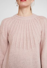 Kobieta w jasnoróżowym swetrze z dzianiny o prążkowanym wzorze i opadających ramionach, uzupełniona przezroczystymi kolczykami w kształcie łzy.