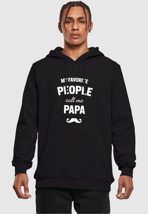 Melnā kapučjaka, kas izgatavota no kokvilnas, ar priekšējo kabatu un baltu tekstu "MANI MĪĻĀKIE CILVĒKI sauc mani par PAPA," ar nelielu ūsām grafikā.