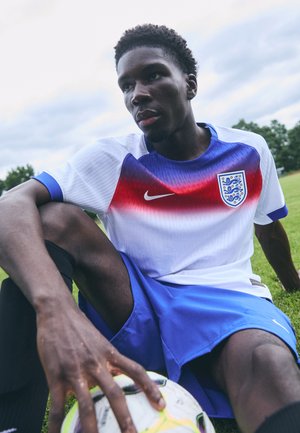Maglia da calcio bianca e blu con accenti rossi in gradiente ed emblema. Pantaloni neri e pallone sull'erba. Focus sulla posizione delle mani.
