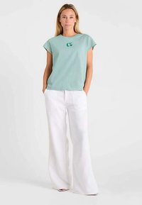 Hellgrünes Baumwoll-T-Shirt mit Rundhalsausschnitt und kurzen Ärmeln, ausgestattet mit einem kleinen grünen Logo, kombiniert mit weißen weit geschnittenen Hosen.