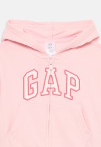 Haut à capuche pour bébé de couleur rose clair avec fermeture éclair avant, poche kangourou et lettres "GAP" en blanc bordées de broderie rose foncé.