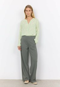Blouse verte clair à motif floral avec un col en V et un détail de boutons, associée à un pantalon gris taille haute et ample, et des ballerines beiges.
