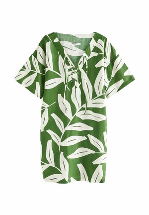 Personne portant une robe verte et blanche à motif de feuilles, debout avec un pied dans l'eau peu profonde à la plage.