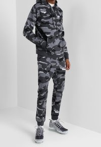 Grå hoodie och sweatpants i camouflage-mönster, gjorda av mjukt material, med en framficka och elastiska muddar, tillsammans med grå träningsskor.