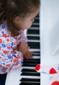 Un enfant portant une chemise à motifs floraux joue du piano blanc, concentré sur les touches noires et blanches. Une chaussette rouge et blanche est posée sur la surface du piano.