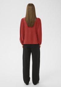 Femme aux cheveux longs et raides de dos, portant un chemisier rouge à manches longues et un pantalon large noir sur un fond uni.